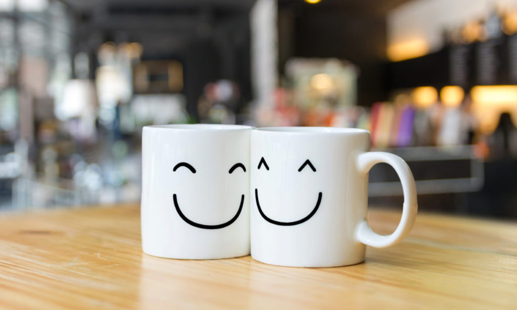 Two white coffee mugs with smiley faces on them sit on a light colored wood table