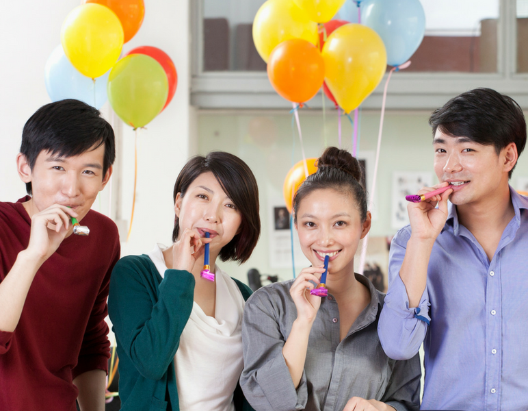 Colleagues hold colorful party noise makers to their mouths while smiling at the camera in front of balloons