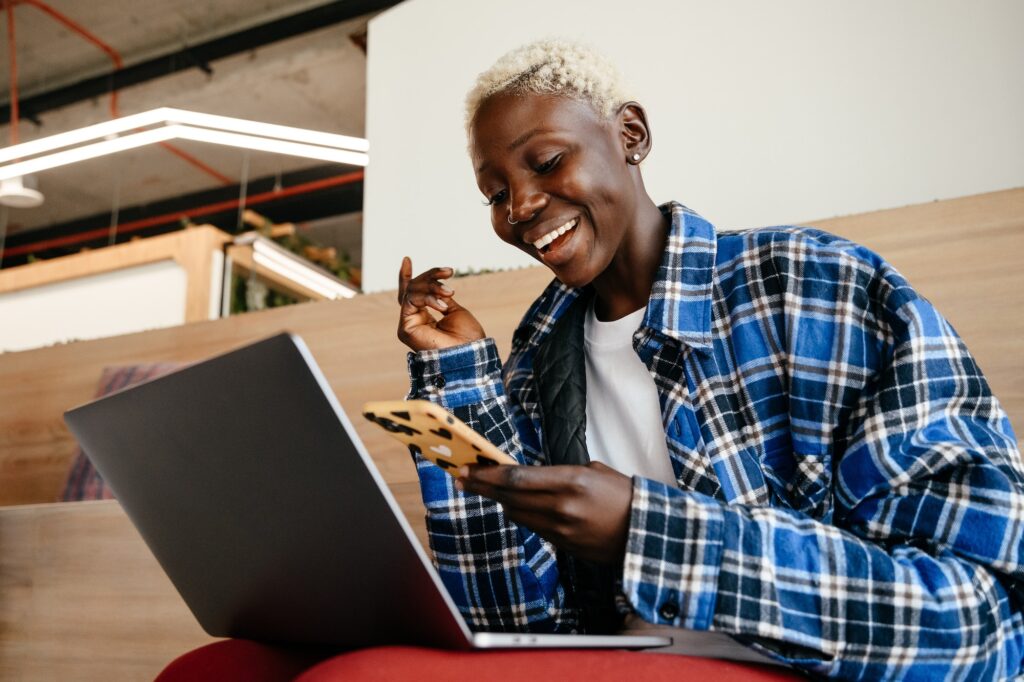 Business owner sitting on a bench in a co-working space, looking at their computer and phone while smiling.